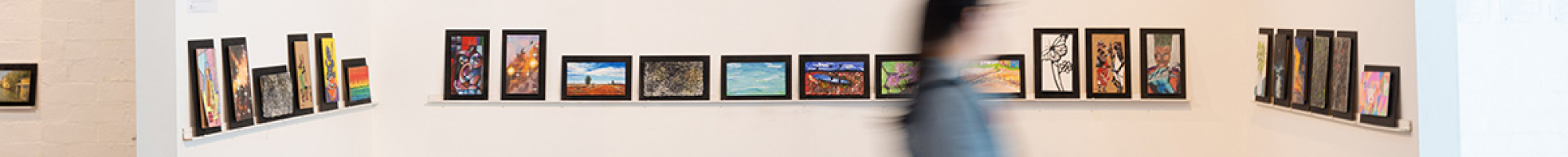 A blurred figure walks in front of a wall of 9 by 5 artworks.