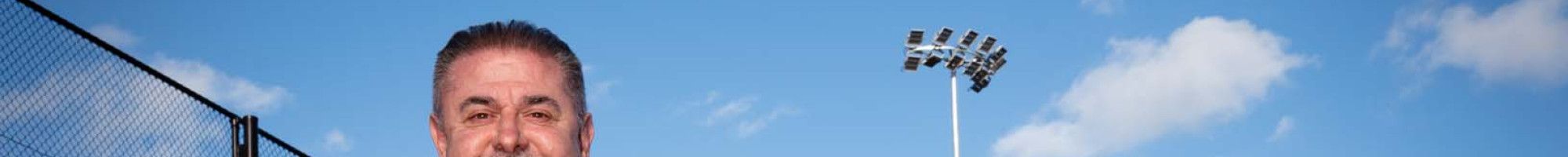 Man in a dark suit stands on a sports ground under a large light tower