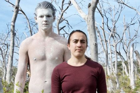 Two performers, one covered in white body paint, in front of bare trees
