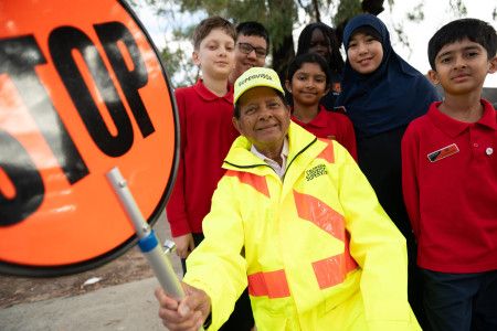 A smiling man dressed in high vis holding a stop sign while surrounded by smiling school children