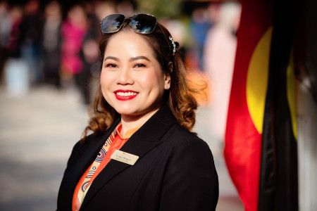 A smiling woman in a black jacket stands side on and smiles to the camera with an Aboriginal flag in the background.