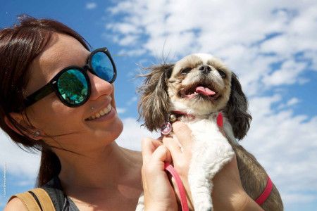 A woman wearing sunglasses holds a brown and white dog up next to her face in front of a cloudy blue sky.
