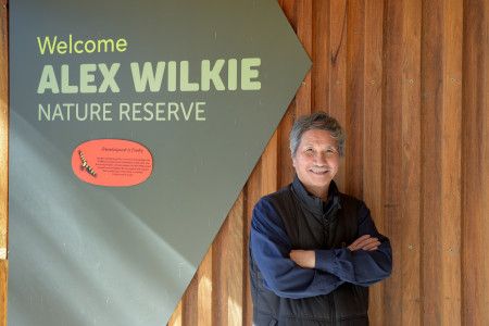 A smiling man with his arms crossed next to a sign that reads Welcome Alex Wilkie Nature Reserve