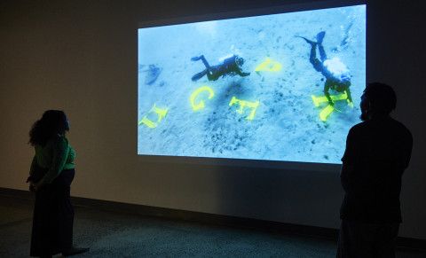 People standing in front of a television screen 'People underwater collecting letters'