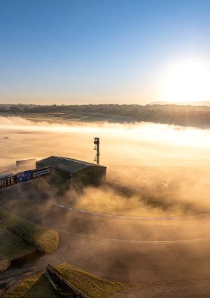 Looking down over the home straight of a racing track that is covered in mist as the sun rises.